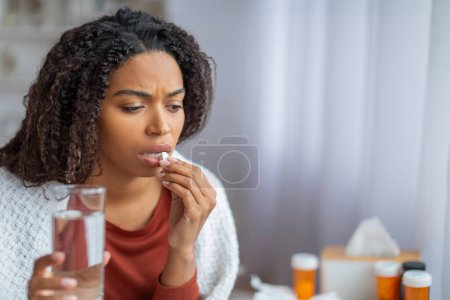 Téléchargez les photos : Jeune femme noire malade prenant une pilule avec un verre d'eau à la maison, femme afro-américaine souffrant de problèmes de santé, assise sur un canapé recouvert de plaid, reflétant un moment d'autosoin à la maison, espace de copie - en image libre de droit