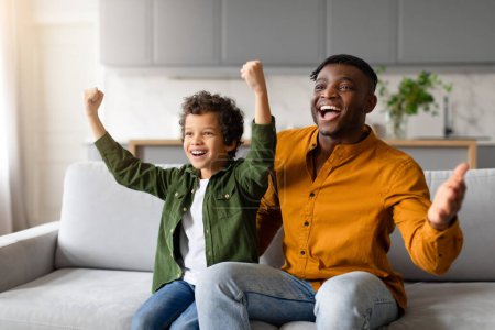 Téléchargez les photos : Joyeux père africain et son fils aux cheveux bouclés lèvent les bras avec enthousiasme dans le geste de victoire tout en regardant le match de football, assis sur le canapé à la maison - en image libre de droit