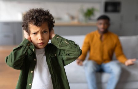Photo for Black child covering his ears with blurred angry father gesturing in the background, depicting family disagreement, selective focus on upset kid - Royalty Free Image