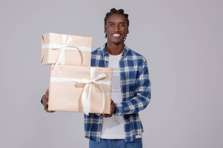Foto de Hora de la celebración. Retrato de tipo negro feliz sosteniendo pila de cajas de regalo aisladas sobre fondo gris en el estudio, hombre afroamericano emocionado de pie con montón de regalos envueltos en papel artesanal - Imagen libre de derechos