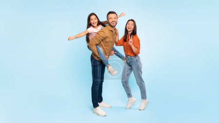 Photo for Fun family time. Happy dad carries his laughing daughter while mom gives a peace sign, posing together over blue background. Studio shot of parents and their kid, panorama, copy space - Royalty Free Image