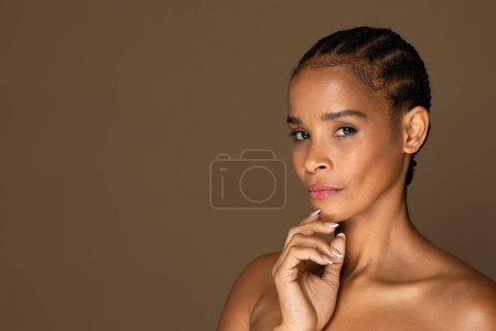 Photo for Reflective black middle aged woman holding her chin in contemplative pose, showcasing natural beauty with braided hair on neutral brown background, free space - Royalty Free Image