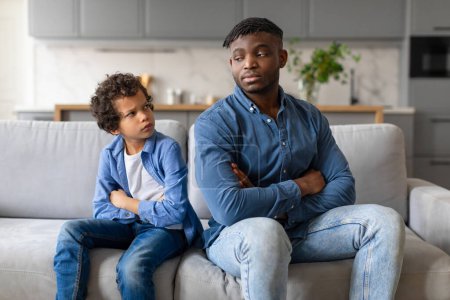 Black father and son sit on the couch with arms crossed, both with serious expressions in moment of family dispute or misunderstanding at home