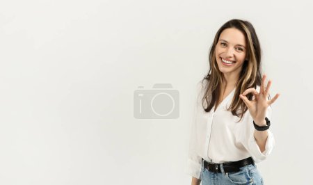 Téléchargez les photos : Jeune femme européenne lumineuse et joyeuse aux cheveux longs qui fait un geste parfait avec sa main, vêtue d'un chemisier blanc, d'un jean denim et d'une smartwatch, sur un fond doux - en image libre de droit