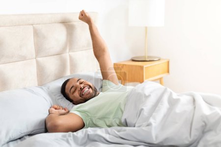 Téléchargez les photos : Heureux homme barbu afro-américain allongé dans son lit et étirant son corps, profitant de bonjour, début de nouvelle journée. Jeune homme noir a bien dormi, intérieur de la maison, espace de copie - en image libre de droit