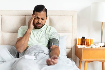 Téléchargez les photos : Homme afro-américain malsain portant un pyjama assis au lit, vérifiant sa tension artérielle avec un tonomètre électronique moderne. Faire un bilan matinal après le réveil ou se sentir malade, copier l'espace - en image libre de droit