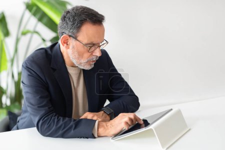 Foto de Hombre de negocios maduro caucásico serio y enfocado con gafas que utilizan intensamente una tableta, reloj inteligente en la muñeca, sentado en una mesa de oficina blanca con exuberantes plantas verdes en el fondo - Imagen libre de derechos