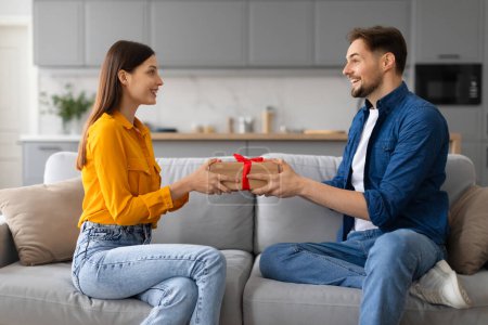 Photo for Young man joyfully gives gift box with red ribbon to smiling woman, capturing moment of happiness and surprise during exchange, birthday or anniversary celebration - Royalty Free Image
