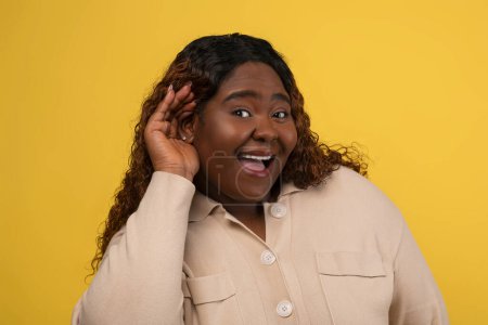 Curious chubby young african american woman in beige shirt with long curly hair holding hand next to ear and grimacing, listening to rumors, gossip, secret. isolated on yellow background, closeup