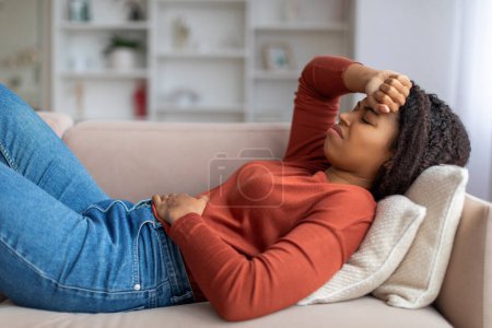 Junge schwarze Frau mit Magenbeschwerden, wenn sie zu Hause auf der Couch liegt, kranke Dame, die Stirn berührt, kranke afrikanisch-amerikanische Dame, die Bauchschmerzen hat, vor Schmerzen Stirnrunzeln, Seitenansicht