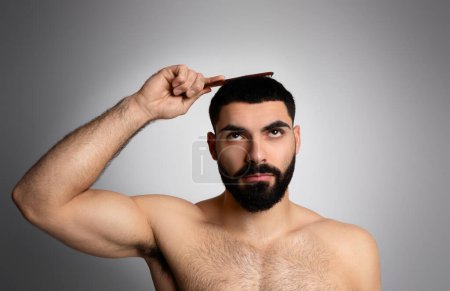 Handsome brunette bearded middle eastern man brushing his thick hair with brush, looking up above his head, posing shirtless isolated on grey studio background. Hair loss, alopecia for men