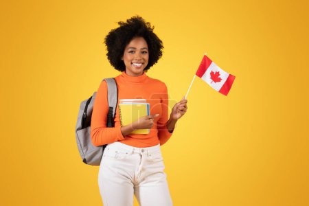 Photo for Smiling young woman with curly hair, dressed in an orange sweater and white jeans, holding a Canadian flag and notebooks, with a backpack, standing against a yellow background - Royalty Free Image