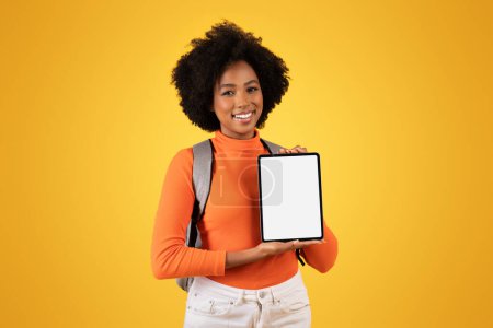 Téléchargez les photos : Amicale jeune femme avec une coiffure naturelle présente un écran de tablette vierge avec un sourire, portant un pull orange et un jean blanc, accessoirisé avec un sac à dos gris sur fond jaune - en image libre de droit
