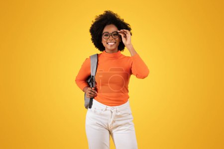 Téléchargez les photos : Une jeune universitaire confiante ajuste ses lunettes élégantes, rayonnantes d'un sourire, vêtue d'un pull orange, d'un jean blanc, avec une sangle grise au-dessus de son épaule sur fond jaune - en image libre de droit