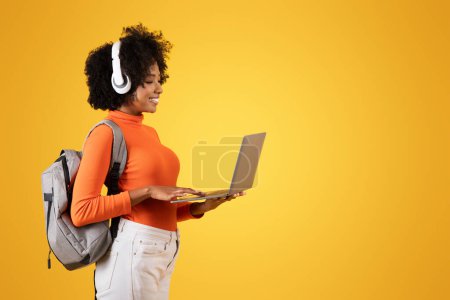 Photo for Focused young woman with curly hair and white headphones, using a laptop while wearing an orange turtleneck and white pants, with a grey backpack, against a yellow background - Royalty Free Image
