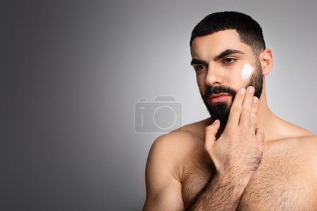 Facial skincare. Middle eastern shirtless man applying moisturizing cream on face, looking at blank free space, isolated on grey studio background. Male beauty and skin care concept
