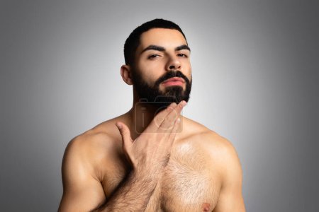 Foto de Guapo atlético medio desnudo joven hombre de Oriente Medio tocando su barba y mirando a la cámara, posando aislado sobre fondo gris estudio. Cuidado de belleza para hombres - Imagen libre de derechos