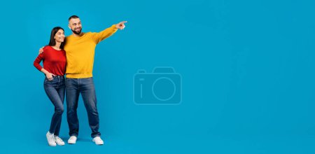 Photo for Check This. Happy Young Couple Pointing Aside At Copy Space, Smiling Millennial Man And Woman Showing Free Place For Your Advertisement Or Offer, Standing Together On Blue Background, Full Length - Royalty Free Image