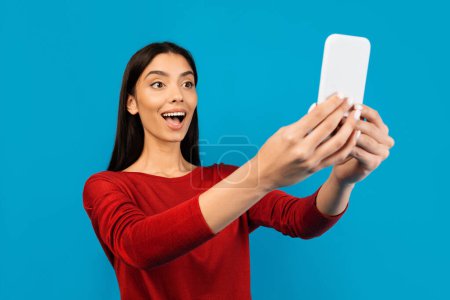 Photo for Great App. Amazed Young Woman Looking At Smartphone Screen With Excitement, Happy Joyful Female Reading Message Or Reacting To Good News, Standing Isolated On Blue Studio Background - Royalty Free Image