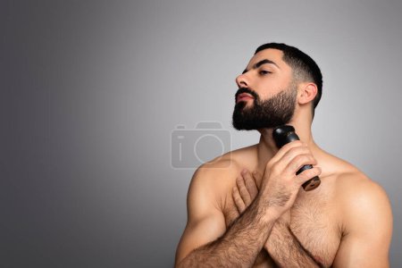 Photo for Beauty, hygiene, shaving, grooming and people concept. Middle eastern young man looking at copy space and shaving beard with trimmer or electric shaver, isolated on grey background - Royalty Free Image