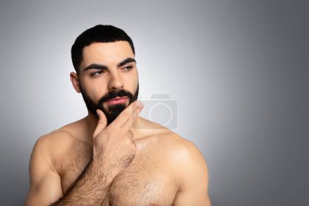 Tips for healthy beard concept. Pensive sporty naked brunette middle eastern young man touching his thick beard and looking at copy space, isolated on grey studio background, closeup