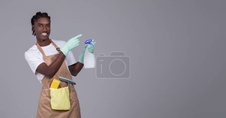 Photo for Young handsome black cleaner guy wearing apron pointing aside at copy space, smiling millennial african american man in gloves, advertising cleaning service, standing against grey studio background - Royalty Free Image