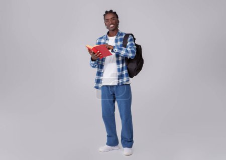 Téléchargez les photos : Nerdy jeune noir étudiant Guy avec livre et sac à dos, sourire bel homme afro-américain dans des lunettes debout isolé sur fond de studio gris, profiter de l'étude et de l'éducation, pleine longueur - en image libre de droit