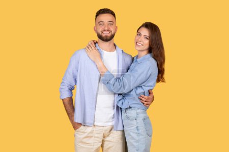 Téléchargez les photos : Joyeux jeune couple aimant en tenue décontractée partager étreinte chaleureuse et sourires joyeux sur fond jaune vibrant, rayonnant de bonheur et de proximité - en image libre de droit