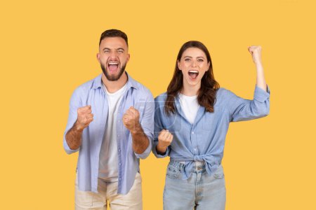 Overjoyed young man and woman with raised fists, celebrating triumph, exuding happiness and success against bright yellow background