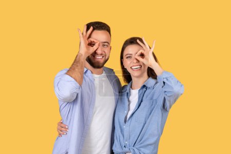 Téléchargez les photos : Joyeux jeune couple faisant des gestes de main ludique comme des lunettes sur leurs yeux, partageant un moment joyeux et léger ensemble sur fond jaune - en image libre de droit