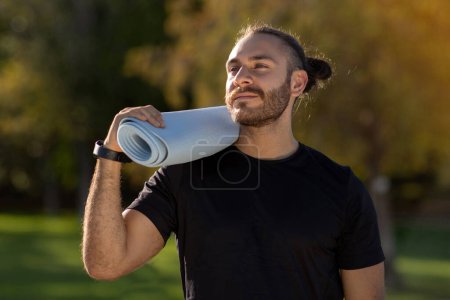 Photo for Young athletic man in black sportswear with smartwatch carrying fitness mat, enjoys summer workout in park outdoor looking away. Concept of wellbeing and sporty lifestyle - Royalty Free Image