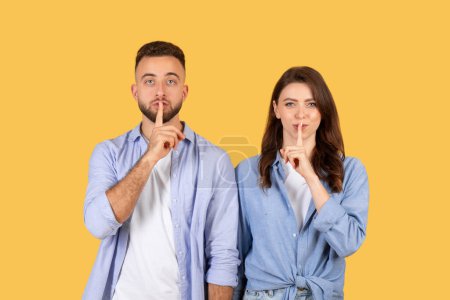 Young man and woman in casual shirts making shush gesture with their fingers on lips, signaling silence, posing against bright yellow background