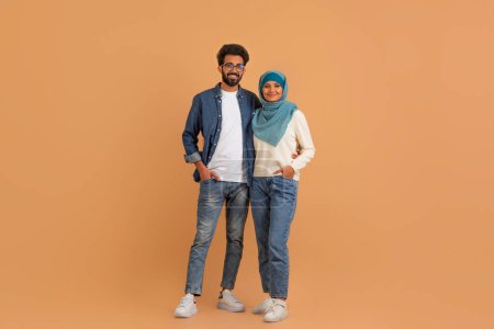 Téléchargez les photos : Portrait de heureux couple musulman moderne debout sur fond de studio beige, Souriant Arabe époux homme et femme dans Hijab embrassant et regardant l'appareil photo, pleine longueur avec espace de copie - en image libre de droit