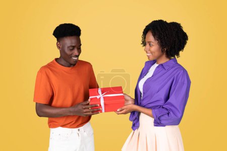 Téléchargez les photos : Un joyeux jeune homme noir heureux dans une chemise orange présente une boîte-cadeau rouge avec un ruban blanc à une femme souriante dans un chemisier violet sur un fond jaune, studio, profiter des vacances - en image libre de droit