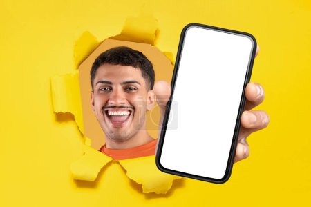 Foto de Hombre risueño con una camisa naranja que atraviesa una pared de papel desgarrada de amarillo, presentando un teléfono inteligente con una pantalla en blanco para contenido personalizable. Gran oferta, anuncio, sitio web de recomendación, aplicación - Imagen libre de derechos