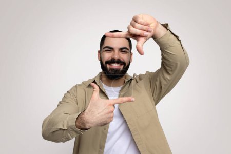 Téléchargez les photos : Jovial et joyeux homme arabe millénaire à la barbe pleine encadre joyeusement son visage d'un geste de main, souriant largement dans une chemise beige sur un fond neutre, studio - en image libre de droit
