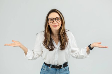 Photo for A young european confused woman with glasses and a friendly expression stands with her arms open, palms up, in a shrugging pose, wearing a white blouse and blue jeans, studio - Royalty Free Image