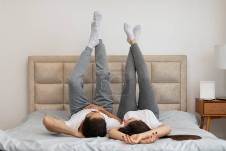 Téléchargez les photos : Couple ludique et détendu jouit moment léger au lit, couché sur le dos avec les jambes en l'air, suggérant confort et facilité - en image libre de droit