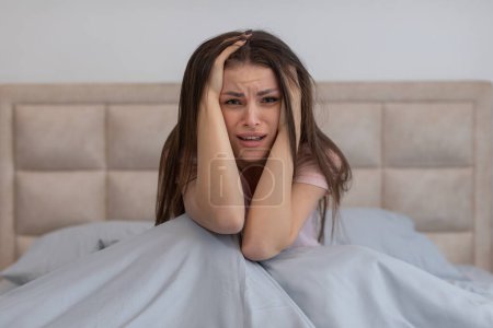 Foto de Mujer joven y ansiosa sentada en la cama con las manos agarradas a la cabeza, mirada de angustia en la cara y fondo de sábanas arrugado - Imagen libre de derechos