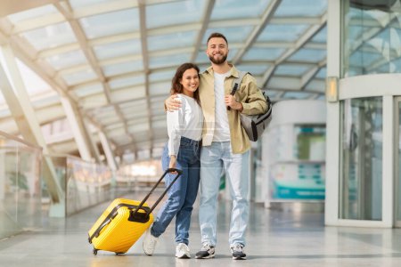 Foto de Listo para el viaje. Feliz joven pareja posando en la terminal del aeropuerto, alegre millennial hombre y mujer de pie con la maleta y sonriendo en la cámara, esperando el vuelo, disfrutando de viajar juntos - Imagen libre de derechos