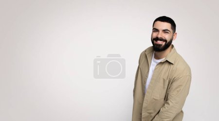 Téléchargez les photos : Un homme jovial avec une barbe et un large sourire se penche légèrement vers la caméra, portant une chemise beige boutonnée sur un t-shirt blanc, sur un fond lumineux spacieux - en image libre de droit