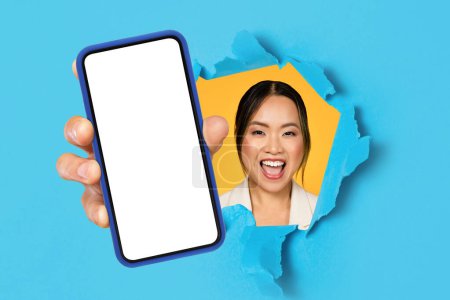 Foto de Sonriendo emocionado joven asiática dama con la boca abierta mostrando el teléfono móvil con pantalla en blanco a través de agujero en papel, fondo de estudio azul. Sitio web, aplicación, recomendación para negocios, trabajo - Imagen libre de derechos