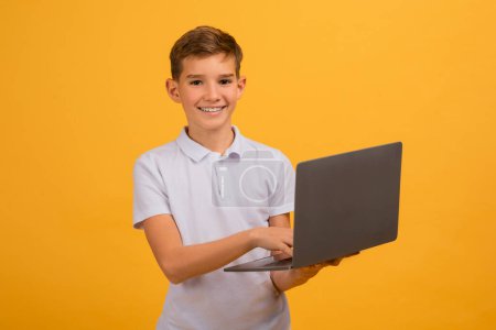 Photo for Portrait Of Smiling Teen Boy Holding Laptop Computer In Hands And Looking At Camera, Happy Teenage Male Kid Enjoying Modern Technologies For Study, Standing On Yellow Studio Background, Copy Space - Royalty Free Image