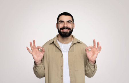 Téléchargez les photos : Un homme barbu millénaire joyeux avec une chemise beige décontractée et un tee-shirt blanc pose avec un sourire confiant, faisant un geste correct avec les deux mains sur un fond clair - en image libre de droit