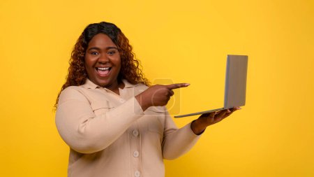 Photo for Happy emotional pretty overweight african american woman student pointing at computer laptop in her hand, isolated over yellow studio background, purchases cheap course. Online education - Royalty Free Image