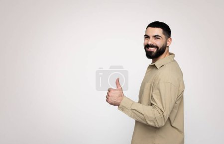 Téléchargez les photos : Profil latéral d'un homme barbu millénaire souriant dans une chemise beige donnant un signe pouce vers le haut, exsudant une ambiance conviviale et accessible, recommander annonce sur un fond clair, studio - en image libre de droit