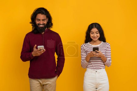 Téléchargez les photos : Homme et femme indiens millénaires utilisant des téléphones cellulaires sur fond de studio jaune, vérifier les médias sociaux, websurf, défilement, lecture de nouvelles, SMS amis. Concept de dépendance au gadget - en image libre de droit