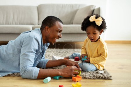 Photo for Beautiful little black girl toddle enjoying time with her daddy at home. Adorable african american father and daughter sitting on floor, playing colorful wooden blocks - Royalty Free Image