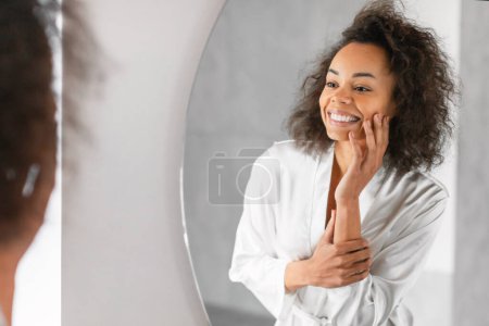 Téléchargez les photos : Jeune femme afro-américaine à la peau impeccable en peignoir de soie se livre à sa routine quotidienne de soins de la peau, touchant doucement son visage devant le miroir de salle de bain à l'intérieur. Teint hydraté doux - en image libre de droit