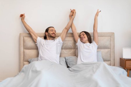 Téléchargez les photos : Couple fraîchement réveillé en t-shirts blancs s'étirant avec les bras levés, saluant le nouveau jour avec joie et énergie dans leur intérieur ensoleillé de chambre - en image libre de droit